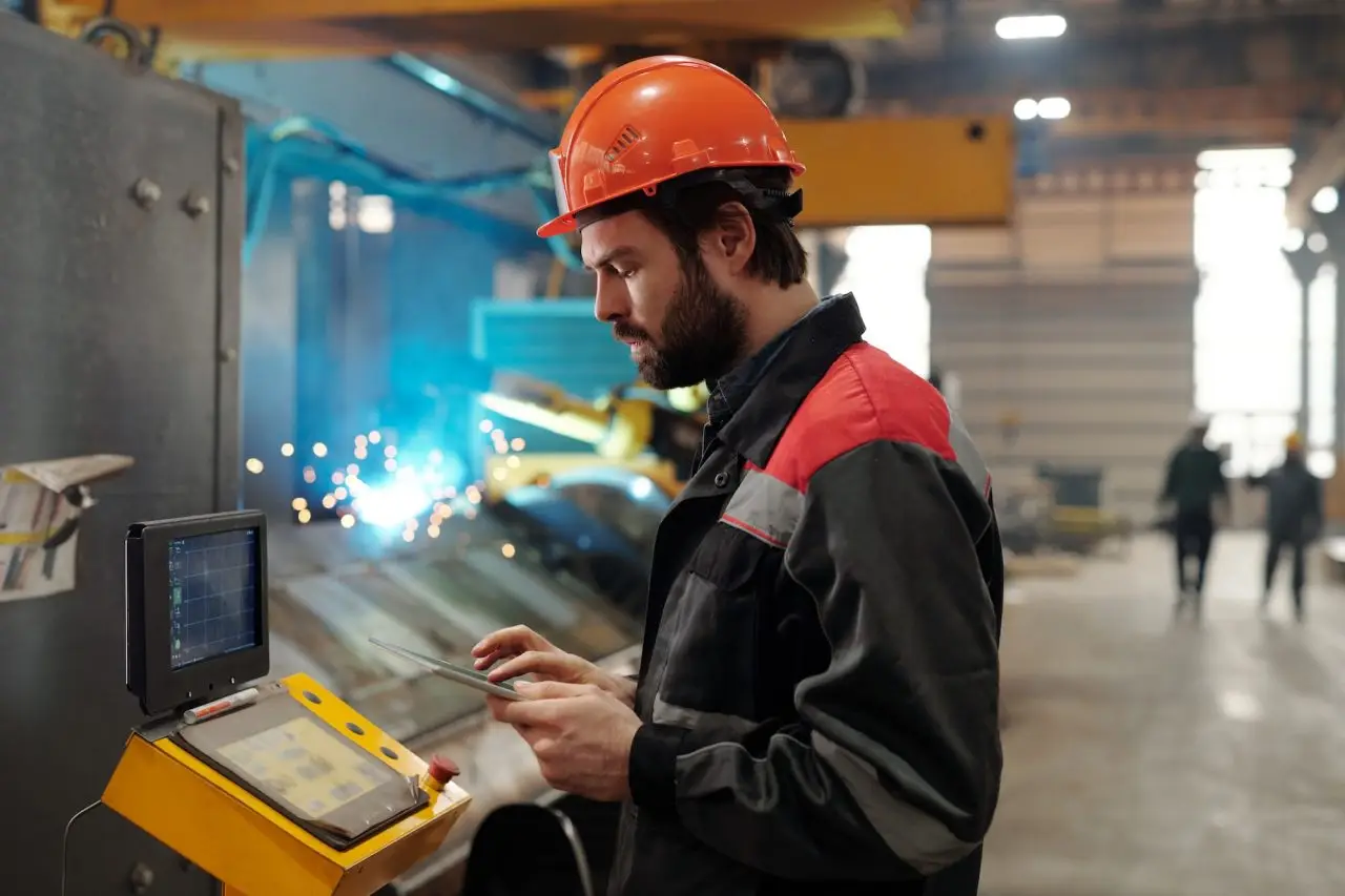 Male factory worker in orange helmet and red-black coveralls operates a welding monitor as blue sparks fly in the background.
