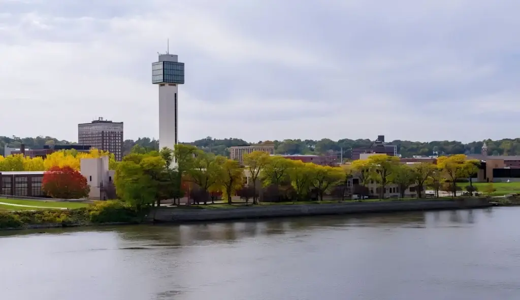 Riverside cityscape with a tall rectangular control-tower building, colorful fall trees, and a calm river in the foreground.