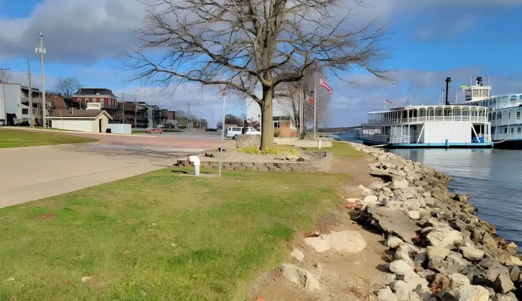 Waterfront park with a large tree, rocky shoreline, and calm water beside a white paddle steamer docked at a docked pier.