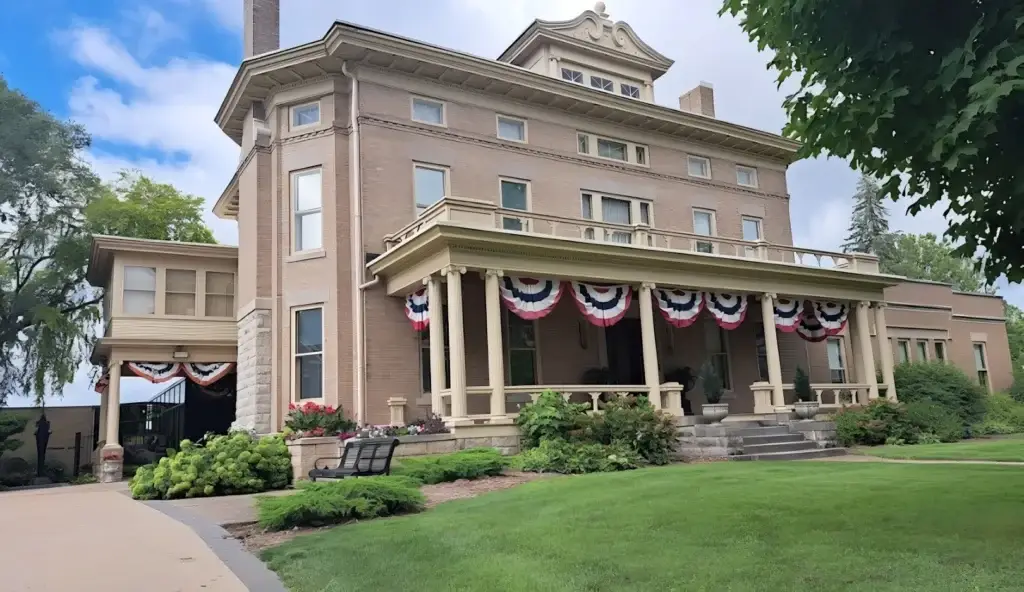 Brick, multi-story mansion with a wraparound porch, cream trim, and red, white, blue bunting along the columns.