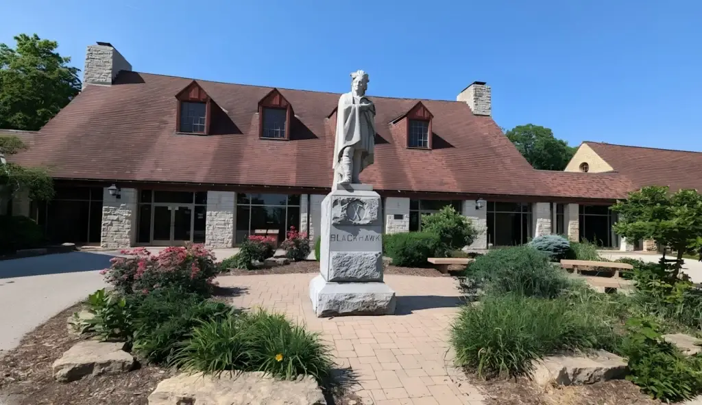 Statue of Black Hawk on a stone pedestal in a courtyard, with a brown-roofed building and landscaped plants behind.