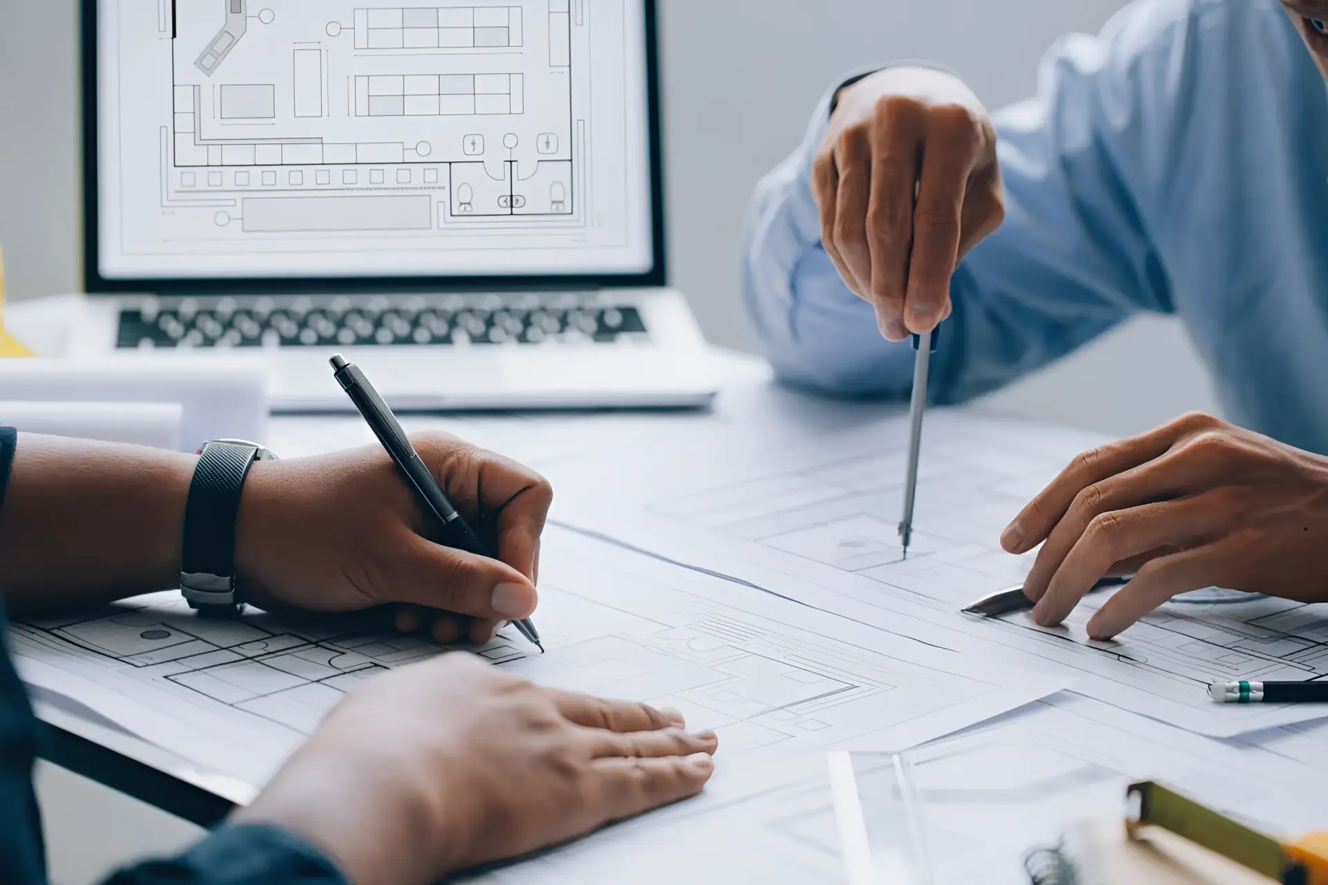 Team of engineers drawing architectural plans on large blueprint sheets with pens, laptop displaying schematics in the background.