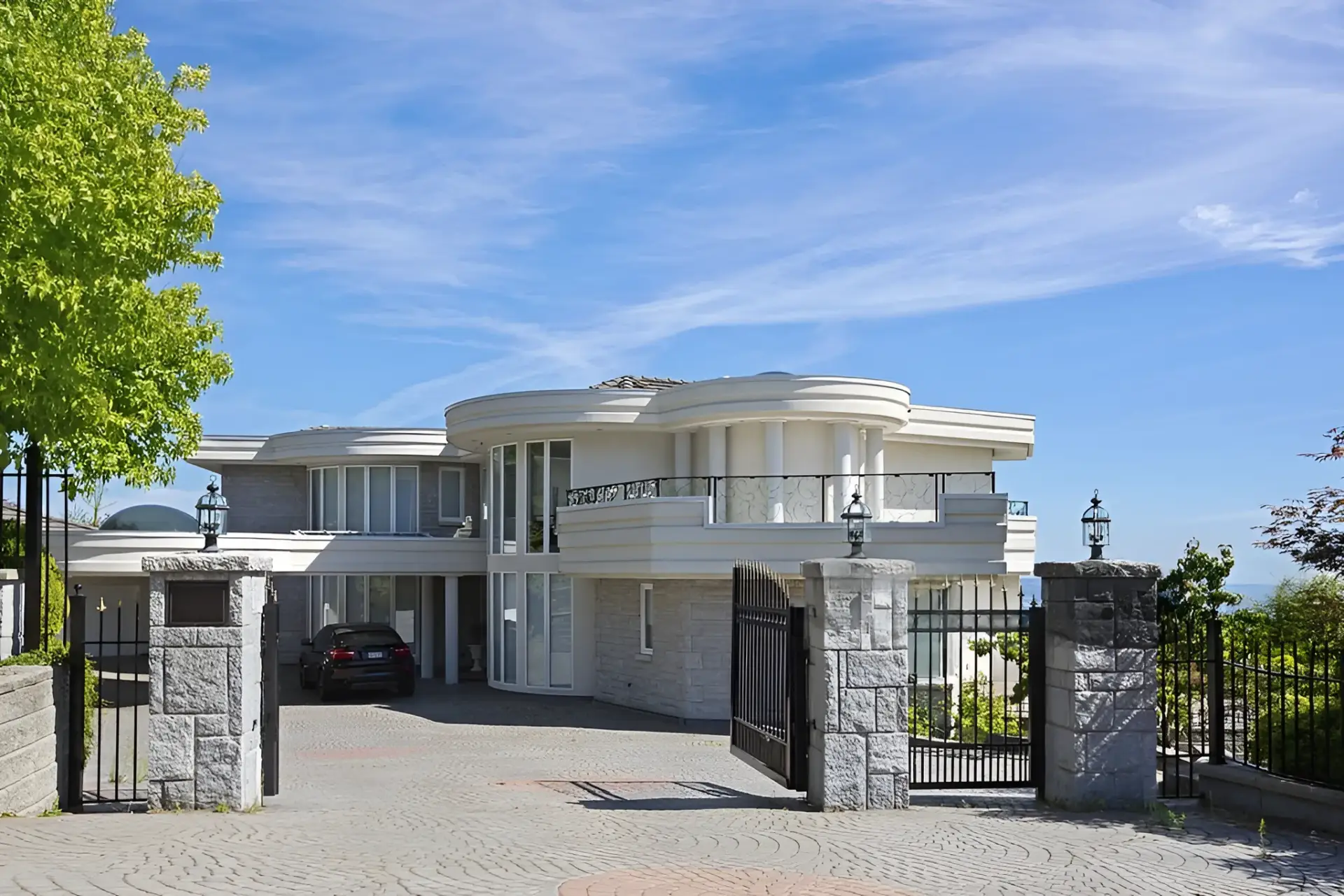 Front view of a modern white villa with curved balconies behind a black iron gate and stone pillars, under a blue sky.