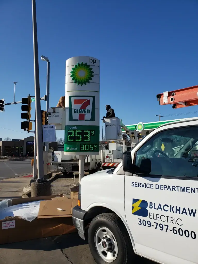 BP gas station sign with 7‑Eleven logo; two workers in bucket lifts work beside the sign, foreground shows a service van labeled 'Blackhawk Electric' and company info.