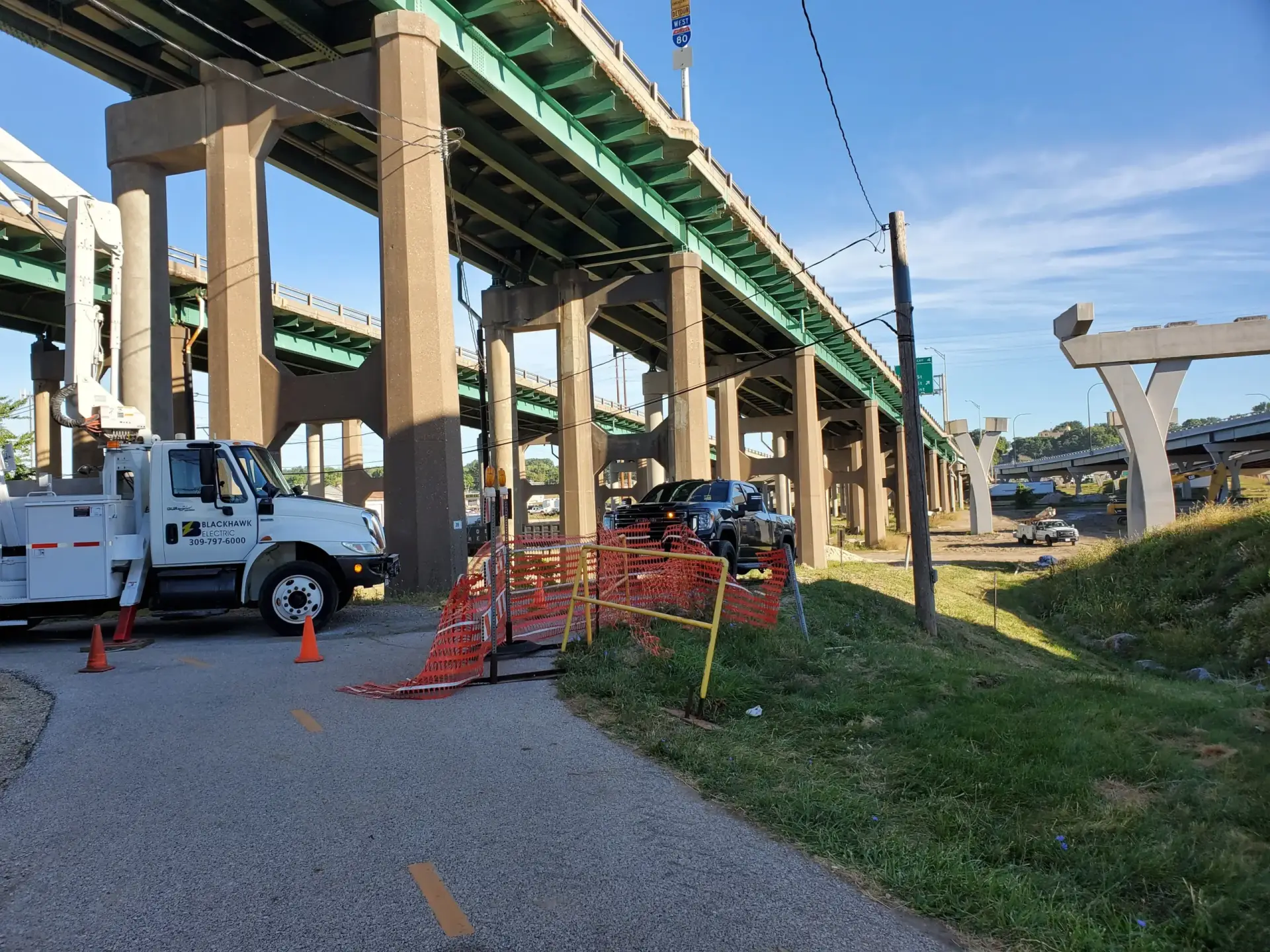 Construction site under a elevated green bridge with concrete supports, orange safety fencing, cones, and work trucks nearby.