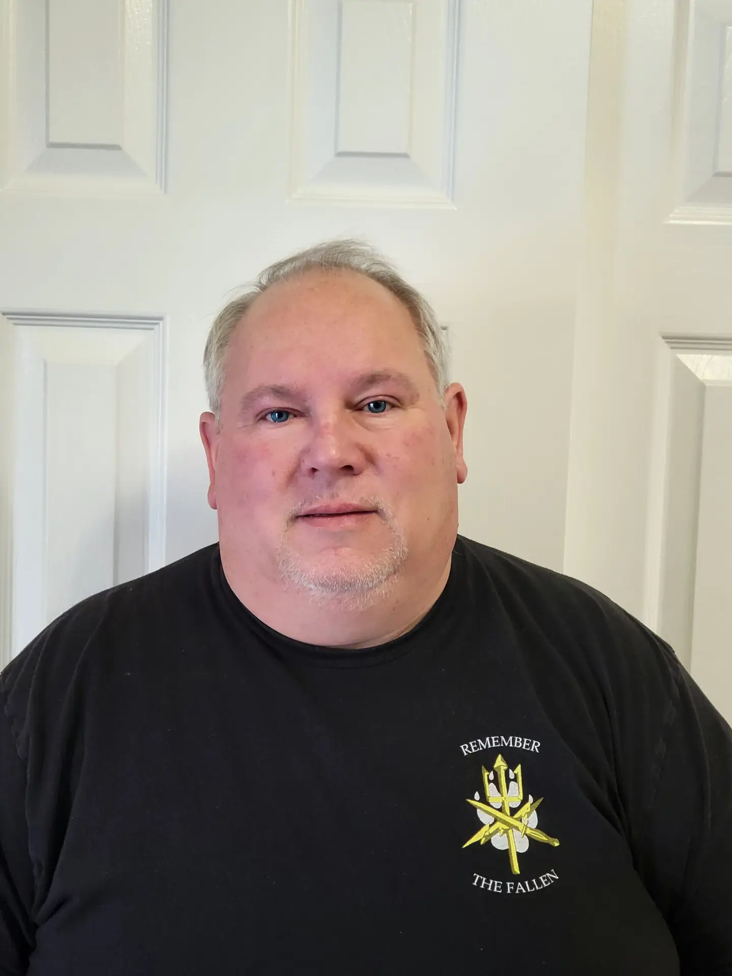 Portrait of a middle-aged man with short gray hair and blue eyes, wearing a black T-shirt with a Remember the Fallen emblem.