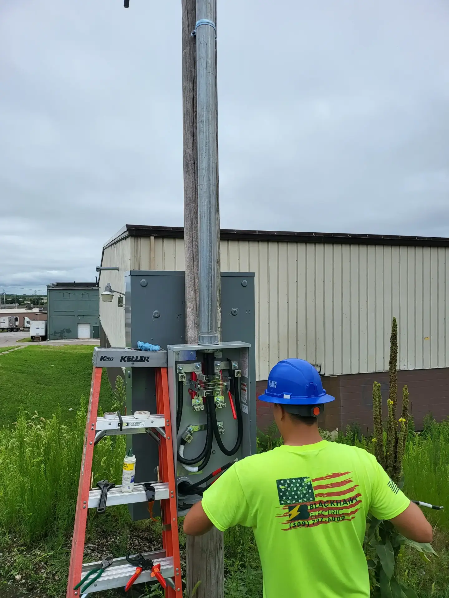 Worker in a neon safety shirt and blue hard hat stands beside a metal electrical cabinet and a pole, performing maintenance work with a ladder nearby.