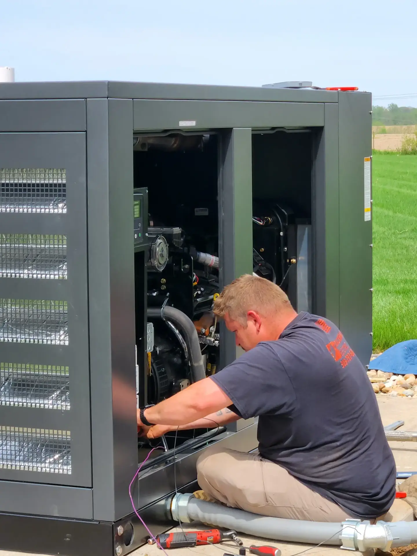 Technician kneels beside an open industrial generator, wires and components exposed outdoors on a grassy area.
