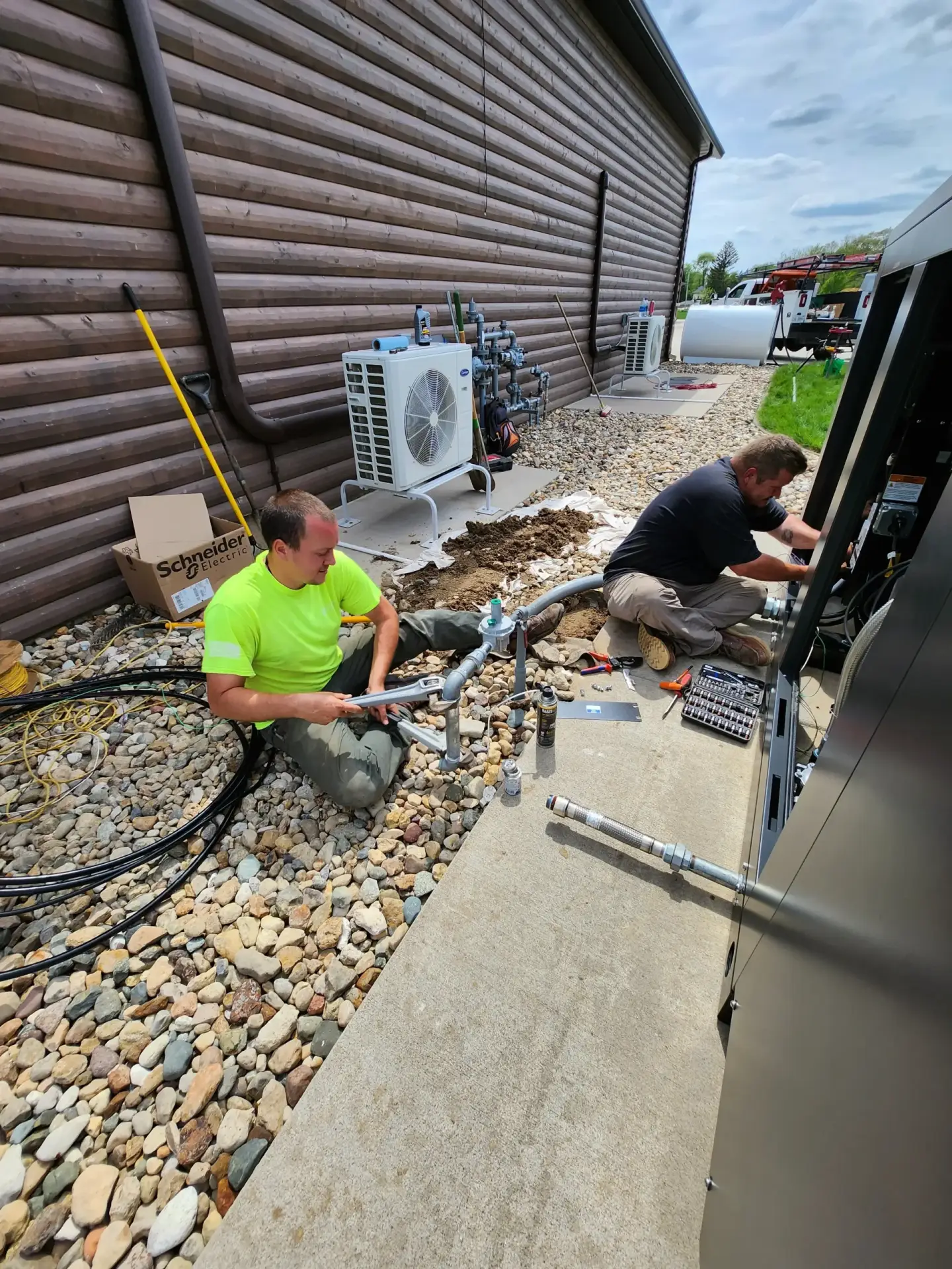 Two technicians in neon shirts work on outdoor HVAC equipment next to a wooden wall, with tools and cables on gravel.
