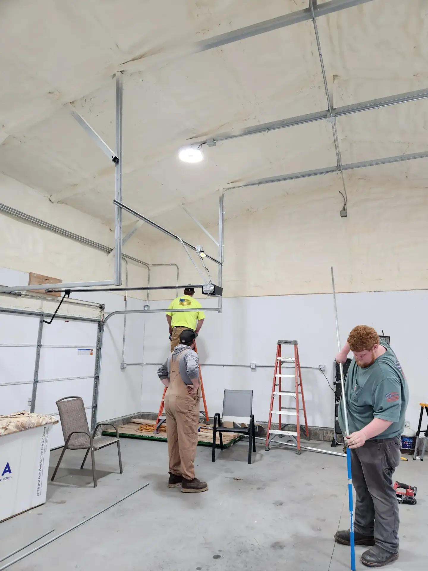 Worker on a ladder installing metal framing in a large unfinished workshop/garage with exposed ceiling beams and conduit