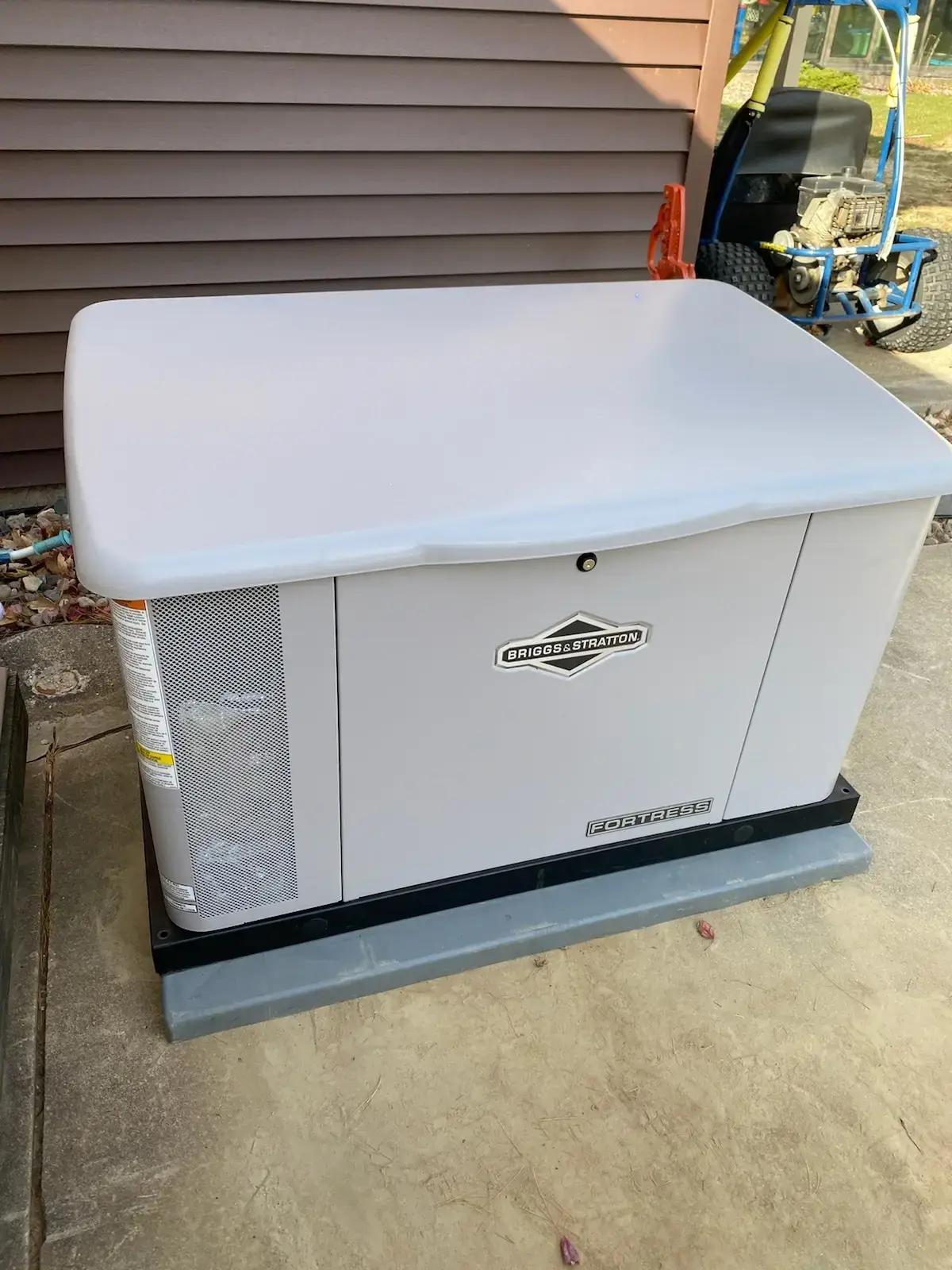 Outdoor Briggs & Stratton Fortress condenser unit on a concrete pad beside a house with brown siding.