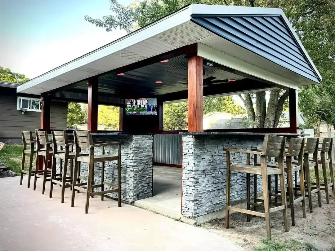 Outdoor bar pavilion with stone columns, wooden bar stools, and a flat-screen TV mounted inside under a gabled roof.