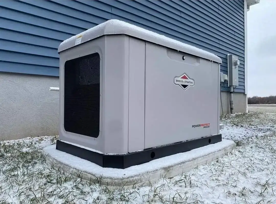 Outdoor air conditioner/condenser unit on a raised concrete pad beside a blue house, light snow resting on top and around the base.
