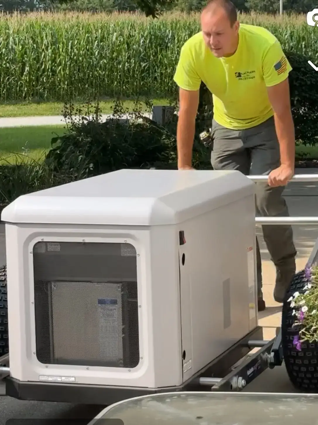 Man in a bright yellow shirt pushing a large white portable generator mounted on a trailer outdoors.