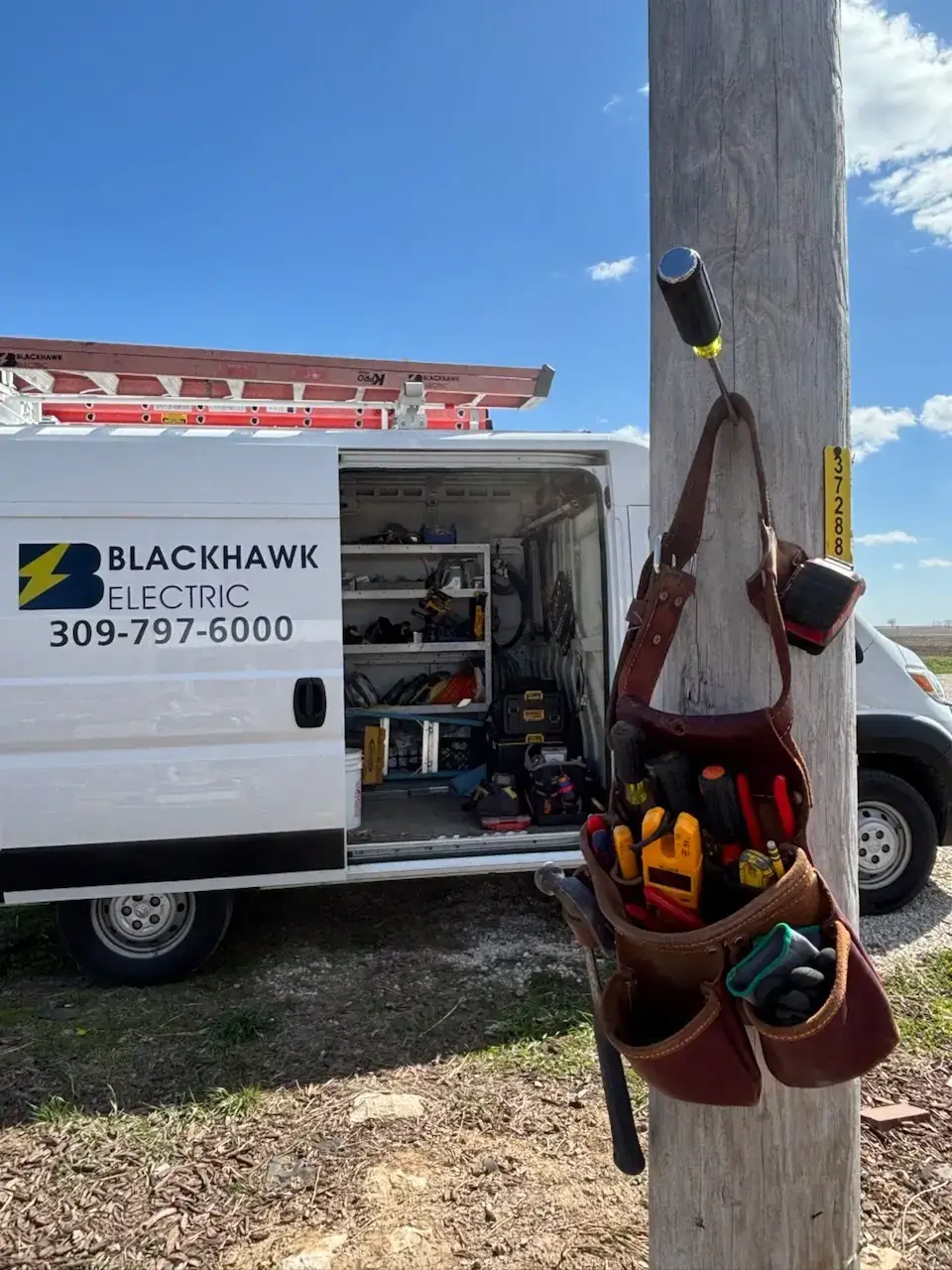 Electrician van with open side door showing shelves of tools and equipment; ladder on top, parked near a utility pole with a tool belt hanging from it.