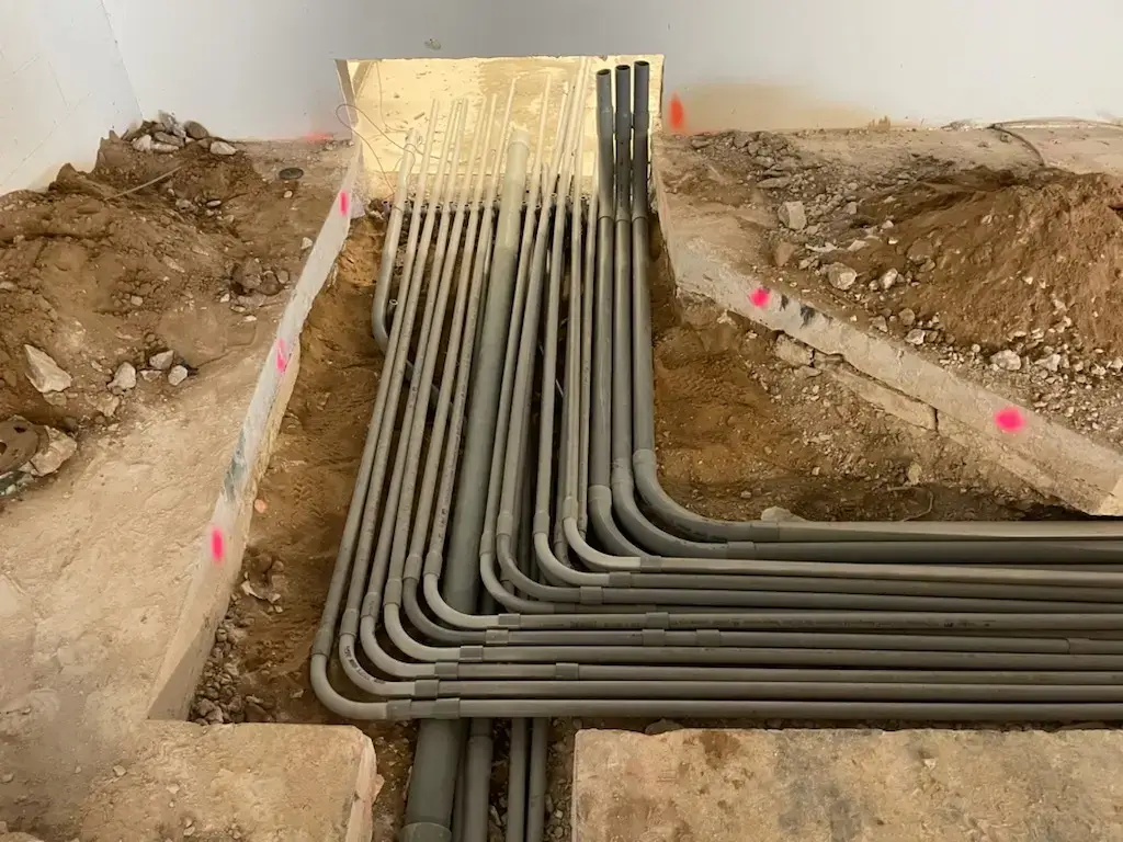 Grey electrical conduits arranged in a trench, bending toward a wall for underfloor installation at a construction site, dirt around them.