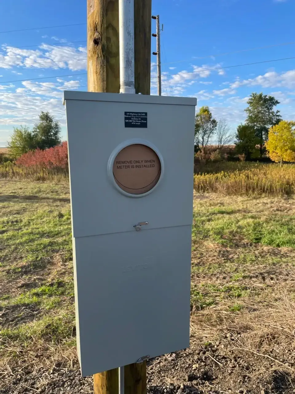Gray utility meter cabinet mounted on a wooden post in a field, with a circular meter window and a small plaque on top edge under a blue sky.