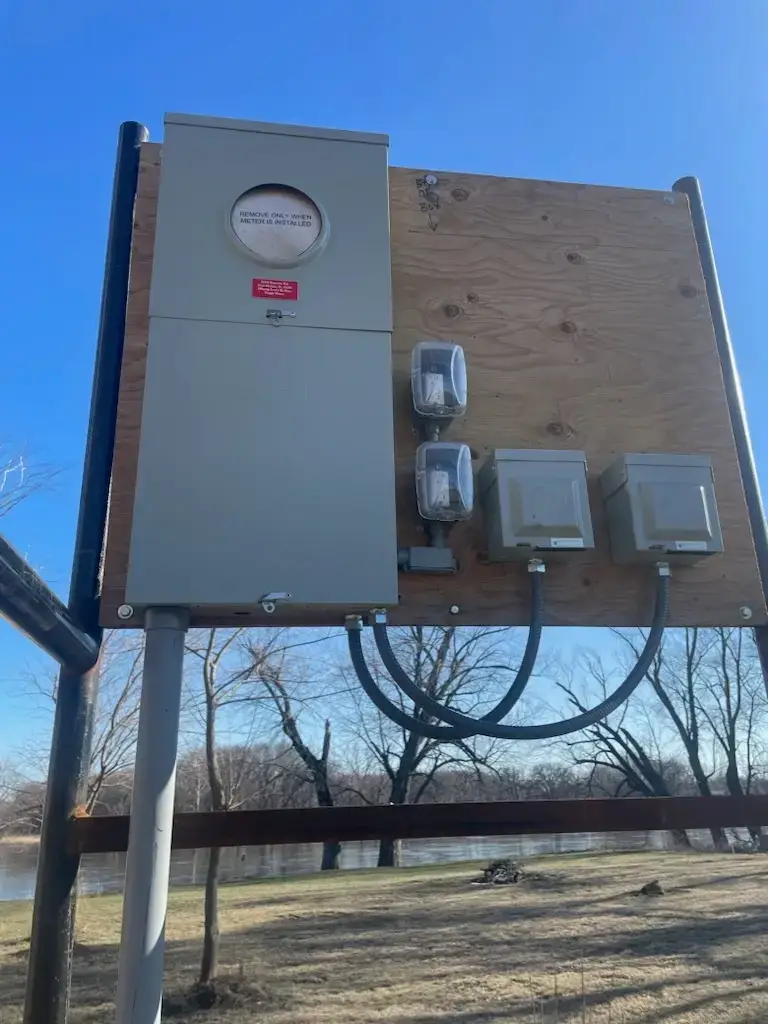 Outdoor electrical service panel mounted on a plywood board with meters and conduits, near a grassy area and trees under a clear blue sky