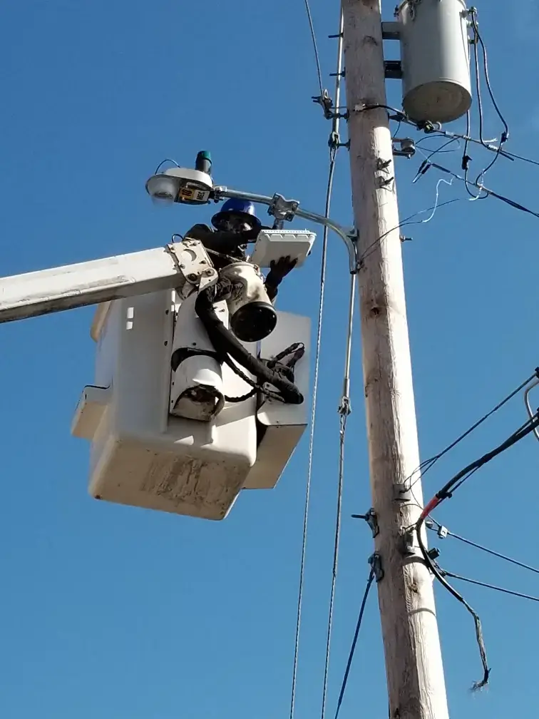 Lineworker in a bucket lift repairing power lines on a wooden utility pole with transformers nearby.
