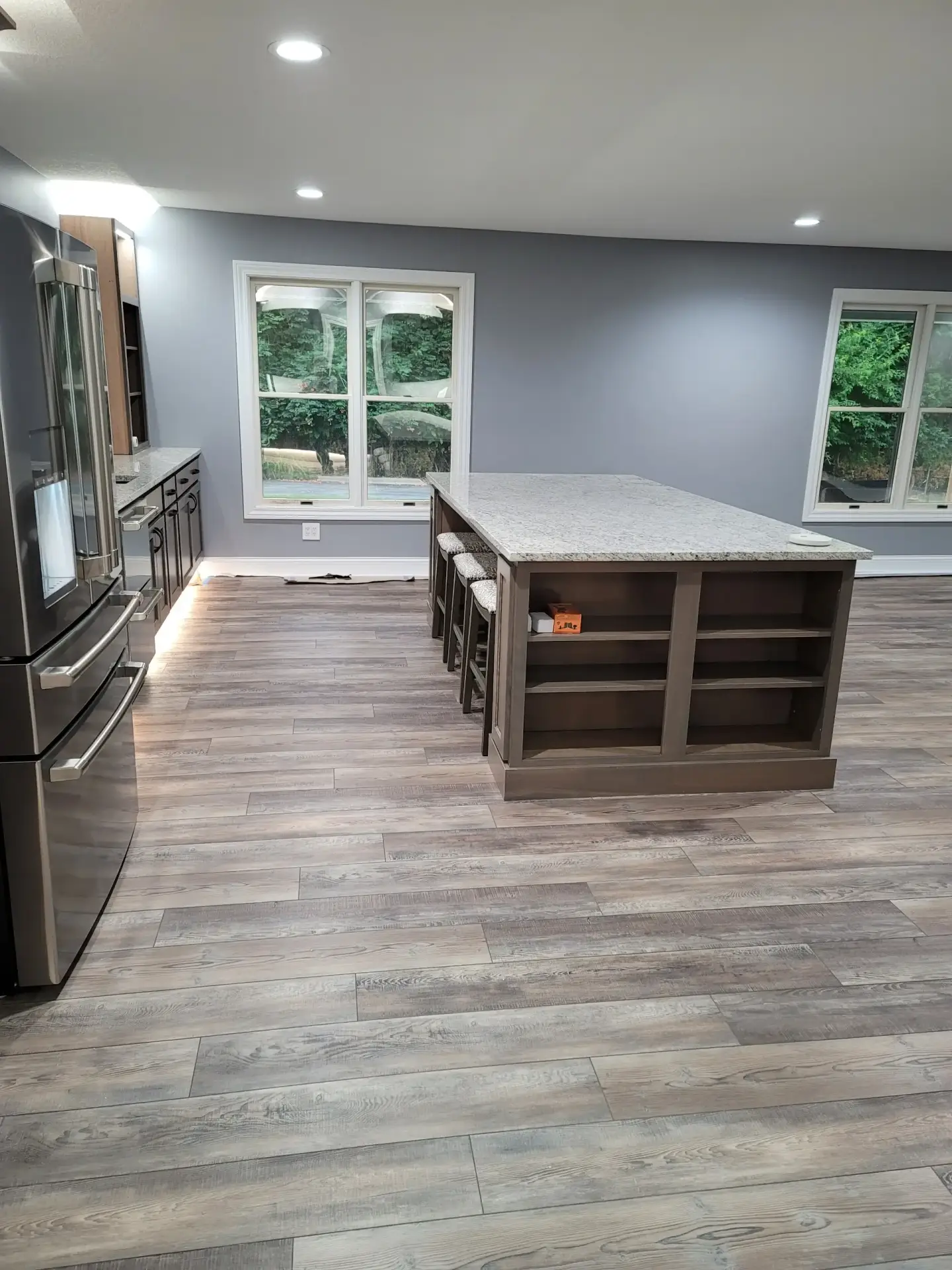 Modern kitchen with a large granite-topped island, bar stools, gray walls, and a stainless steel refrigerator on the left, windowed view outside.