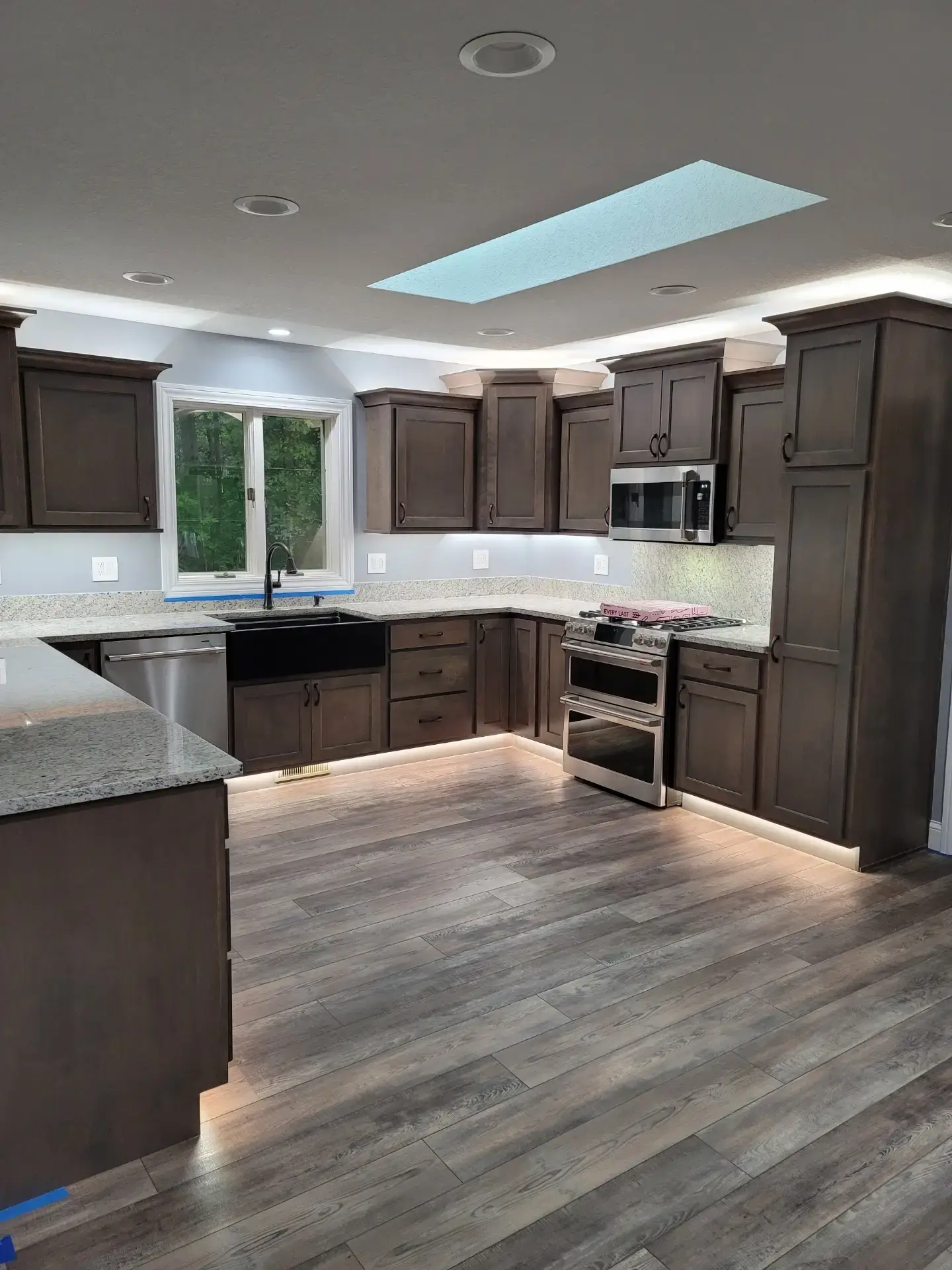 Modern L-shaped kitchen with dark wood cabinets, granite countertops, and stainless steel appliances under soft overhead lighting and a skylight.
